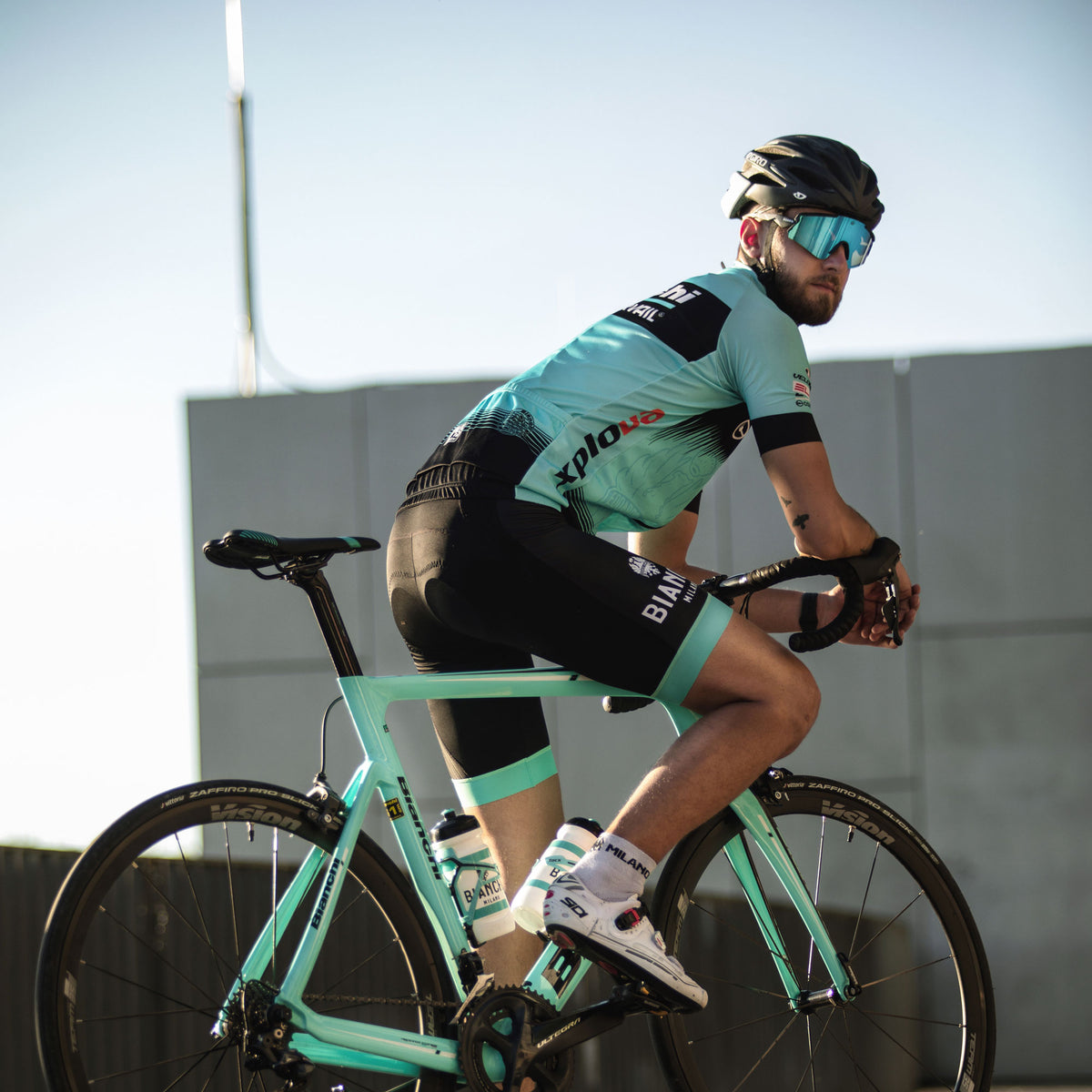 Un ciclista, con abbigliamento verde acqua e nero, pedala all’aperto sotto un cielo sereno su una bicicletta verde acqua, guardandosi alle spalle mentre indossa il modello FALCON di NAKED Optics con protezione UV400.