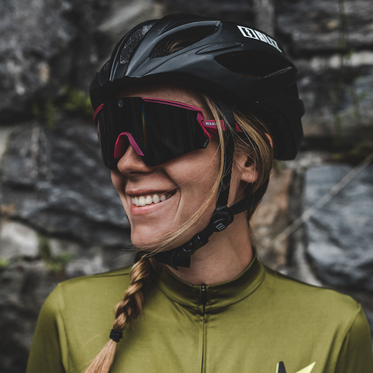 A woman wearing a bike helmet and The FALCON Sportsonnenbrille with UV400 Schutz from NAKED Optics, smiling with a braid, stands in front of a stone wall.