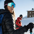 A woman in blue The FALCON Sportsonnenbrille by NAKED Optics with UV400 Schutz and a black jacket holds ski poles; another skier is blurred in the snowy background.