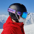 A person in a red jacket, black helmet, and blue ski goggles with Brillenband Befestigung wears an FFP2 Maske secured with the NAKED Optics Maskenclip für FFP2 Maske, standing on a snowy mountain under a blue sky.