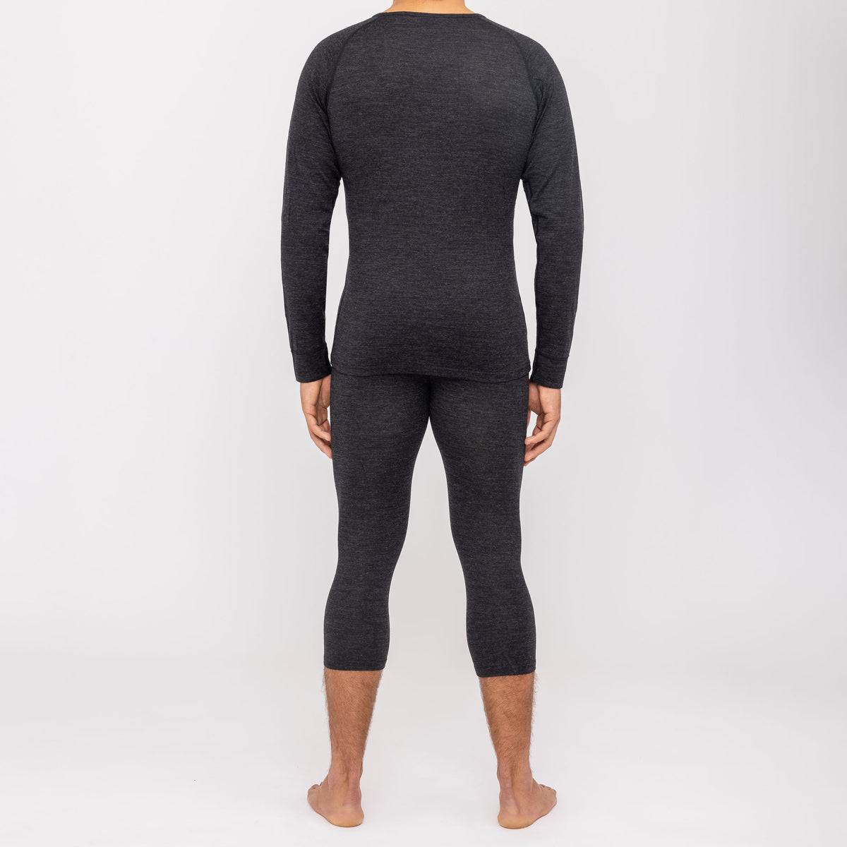 Ein Mann, der die NAKED Optics Thermo-Unterwäsche für Herren (100 % Merino) trägt, steht barfuß in einem dunklen Langarmshirt und passenden Leggings mit dem Rücken zu einem schlichten weißen Hintergrund und unterstreicht so die schlanke Passform dieser nahtlosen Funktionsunterwäsche aus Merinowolle.