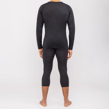 Ein Mann, der die NAKED Optics Thermo-Unterwäsche für Herren (100 % Merino) trägt, steht barfuß in einem dunklen Langarmshirt und passenden Leggings mit dem Rücken zu einem schlichten weißen Hintergrund und unterstreicht so die schlanke Passform dieser nahtlosen Funktionsunterwäsche aus Merinowolle.