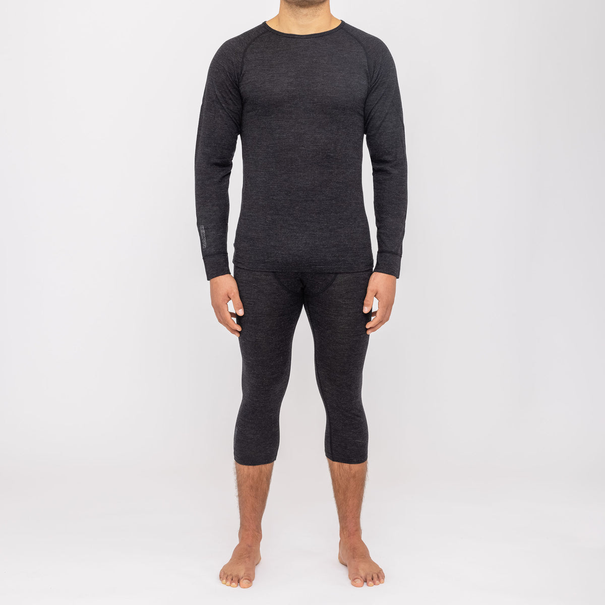 Ein Mann steht barfuß vor einem weißen Hintergrund und trägt ein dunkles Langarmshirt sowie kurze Leggings, die die nahtlose Thermo-Unterwäsche für Herren (100 % Merino) von NAKED Optics zur Geltung bringen. Diese feuchtigkeitsableitende Merino-Funktionswäsche bietet außergewöhnlichen Komfort und Wärme und eignet sich daher ideal zum Kombinieren in den kälteren Monaten.