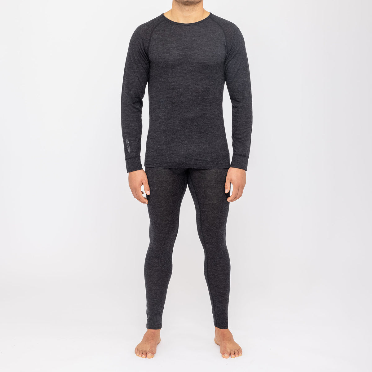 Una persona è in piedi a piedi nudi su uno sfondo bianco e indossa una maglia a maniche lunghe e dei leggings grigio scuro abbinati, della collezione NAKED Optics Thermo-Unterwäsche Herren (100% lana merino).