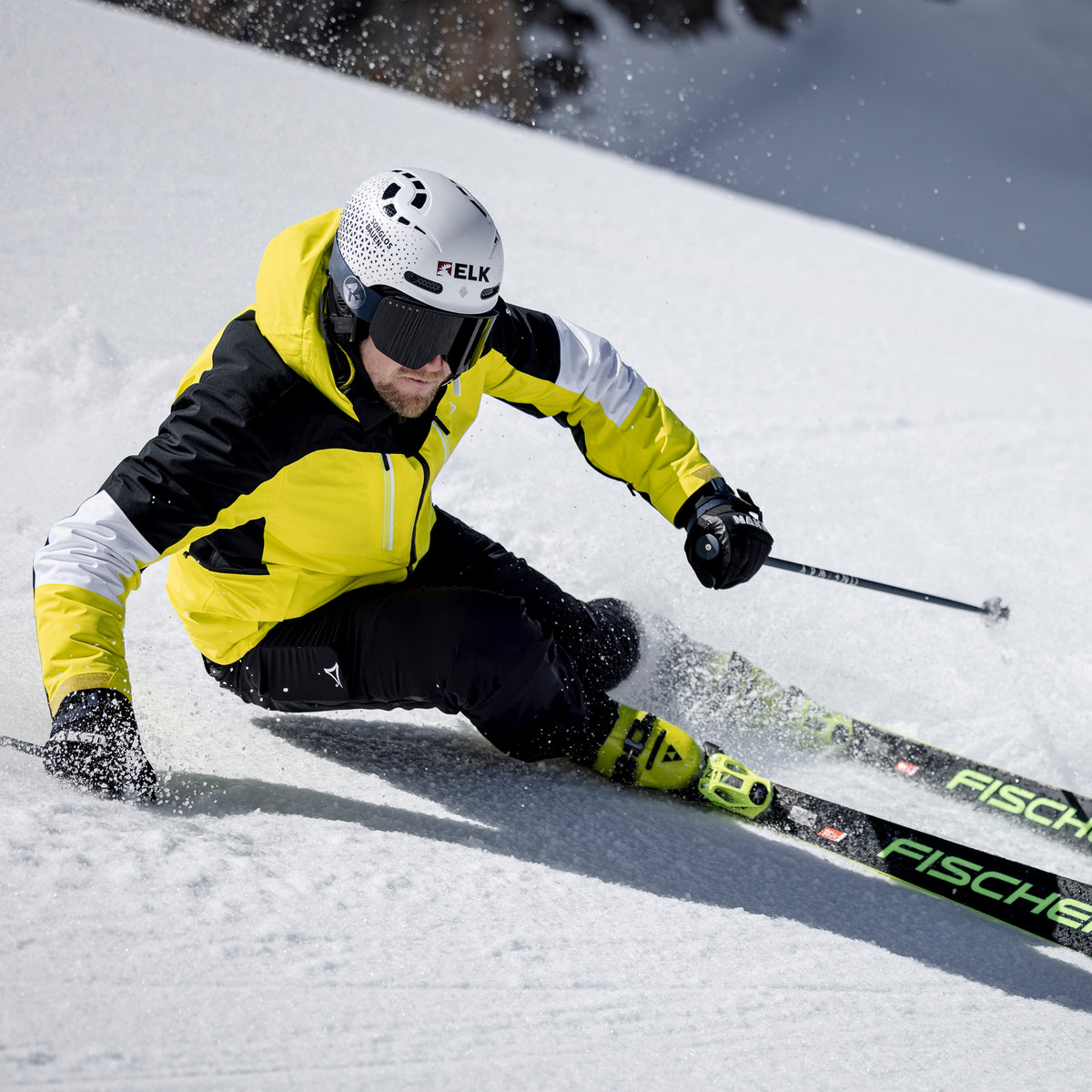 Ein Skifahrer in gelber Jacke und mit Helm, der die NOVA-Skibrille von NAKED Optics mit Anti-Fog-Beschichtung trägt, saust auf schwarzen Fischer-Skiern einen verschneiten Hang hinunter.
