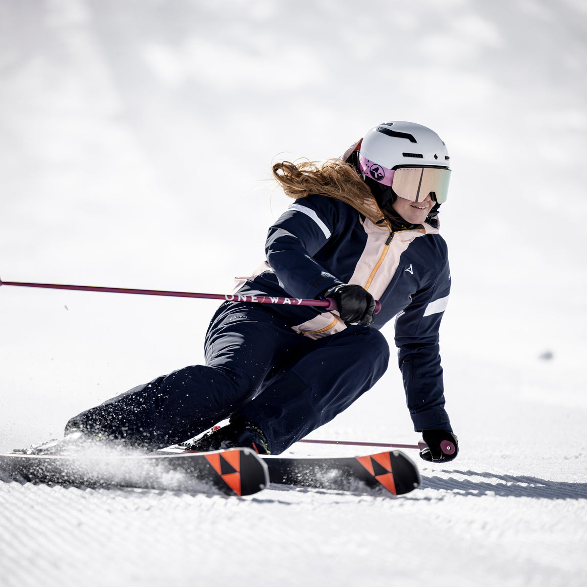 Ein Skifahrer in dunkler Ausrüstung und mit Helm, der die Skibrille „The NOVA“ von NAKED Optics mit Anti-Fog-Beschichtung trägt, macht eine scharfe Kurve auf einem verschneiten Hang und wirbelt dabei Schnee hinter sich auf.