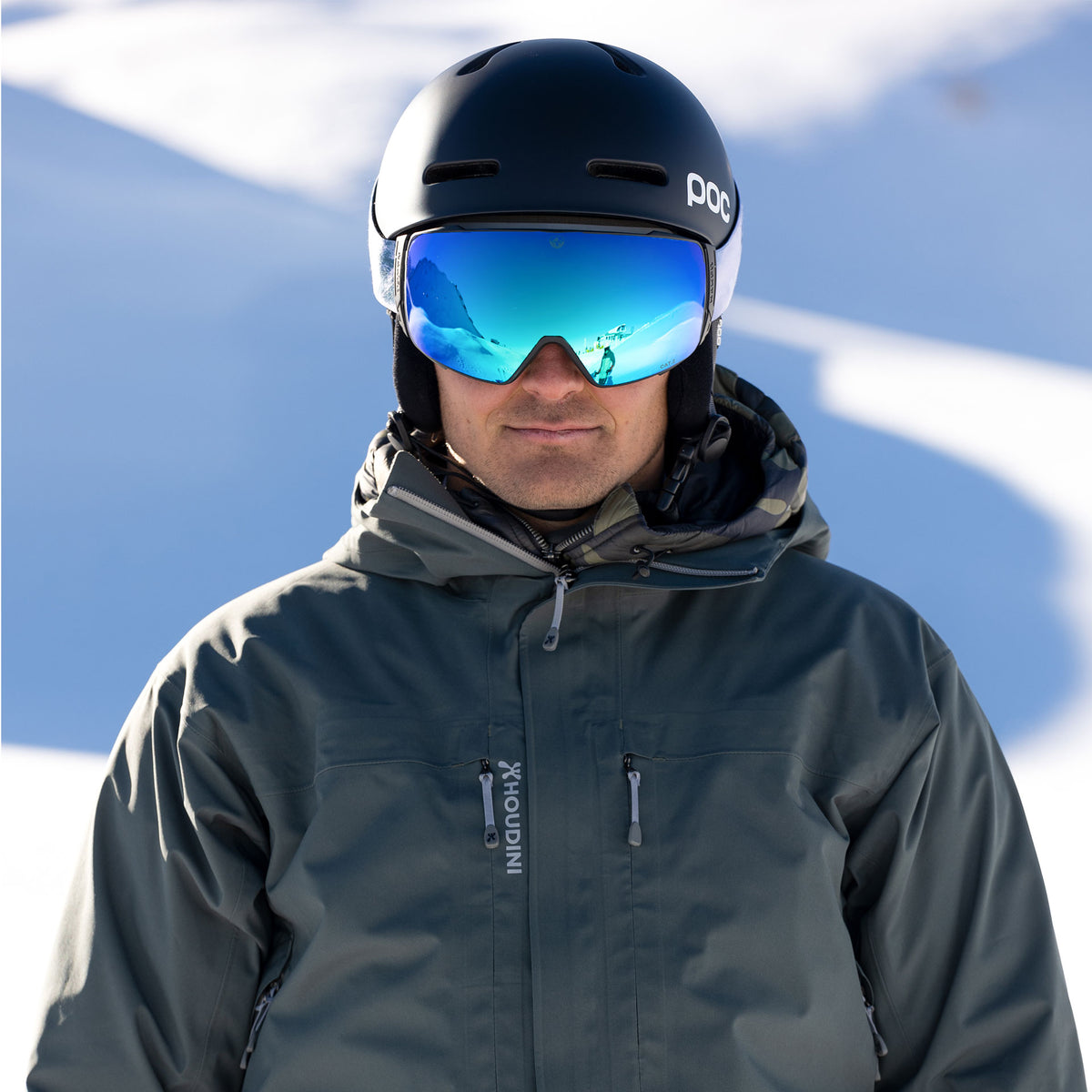 Person in ski gear, helmet, and reflective goggles—The STORM by NAKED Optics with sphärischer Skibrille and Anti-Fog-Beschichtung—standing in a snowy outdoor setting under sunlight.