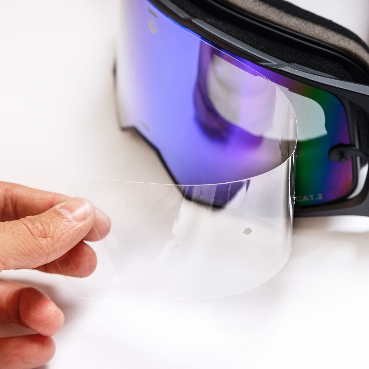 A hand peels a clear protective film from the purple-tinted NAKED Optics Ersatzglas LINE ski and snowboard goggles, showcasing their versatile design and UV-Schutz feature.