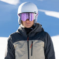 Una persona in tenuta da sci, con indosso un casco bianco e gli occhiali da sci riflettenti NAKED Optics The TROOP Youth Skibrille Jugendliche con rivestimento antiappannamento, si trova in un paesaggio innevato all’aperto.