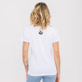 Une femme aux cheveux blonds ondulés, vue de dos avec des tatouages visibles, portant le t-shirt NAKED Optics au logo bicolore, en coton bio, et un jean.