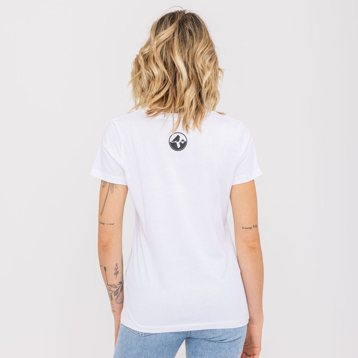 Une femme aux cheveux blonds ondulés, vue de dos avec des tatouages visibles, portant le t-shirt NAKED Optics au logo bicolore, en coton bio, et un jean.