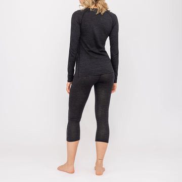 Eine Frau steht barfuß, mit dem Rücken zur Kamera, vor einem weißen Hintergrund und trägt ein schwarzes Langarm-Oberteil sowie kurze Leggings. Dieses stilvolle Set ist die Thermo-Unterwäsche für Damen (100 % Merino) von NAKED Optics, die fachmännisch aus weicher Merinowolle gefertigt wurde und als hochwertige Funktionsunterwäsche für Damen außergewöhnlichen Tragekomfort und Atmungsaktivität bietet.