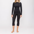 A woman stands barefoot against a plain white background, wearing a long-sleeve black top and black capri leggings from NAKED Optics’ Thermo-Unterwäsche Damen (100% Merino) collection.