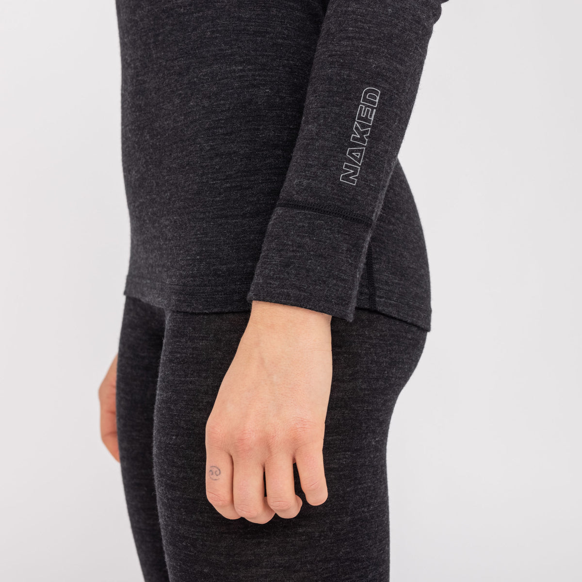 A person is wearing NAKED Optics Thermo-Unterwäsche Damen (100% Merino), featuring a dark long-sleeve top and leggings made from soft Merino wool, with “NAKED” written on the sleeve. Their hand is relaxed.