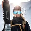 Eine Person in Winterkleidung und mit der reflektierenden FALCON Sportsonnenbrille von NAKED Optics hält vor dem Hintergrund verschneiter Berge und im Sonnenlicht ihre Skier in der Hand und genießt dabei klare Sicht und zuverlässigen UV400-Schutz.