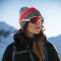 A woman wearing a gray and pink beanie, sunglasses, and a jacket stands outdoors in snowy mountains, her NAKED Optics The FALCON Sportsonnenbrille with UV400 Schutz protecting her eyes from the bright alpine sun.