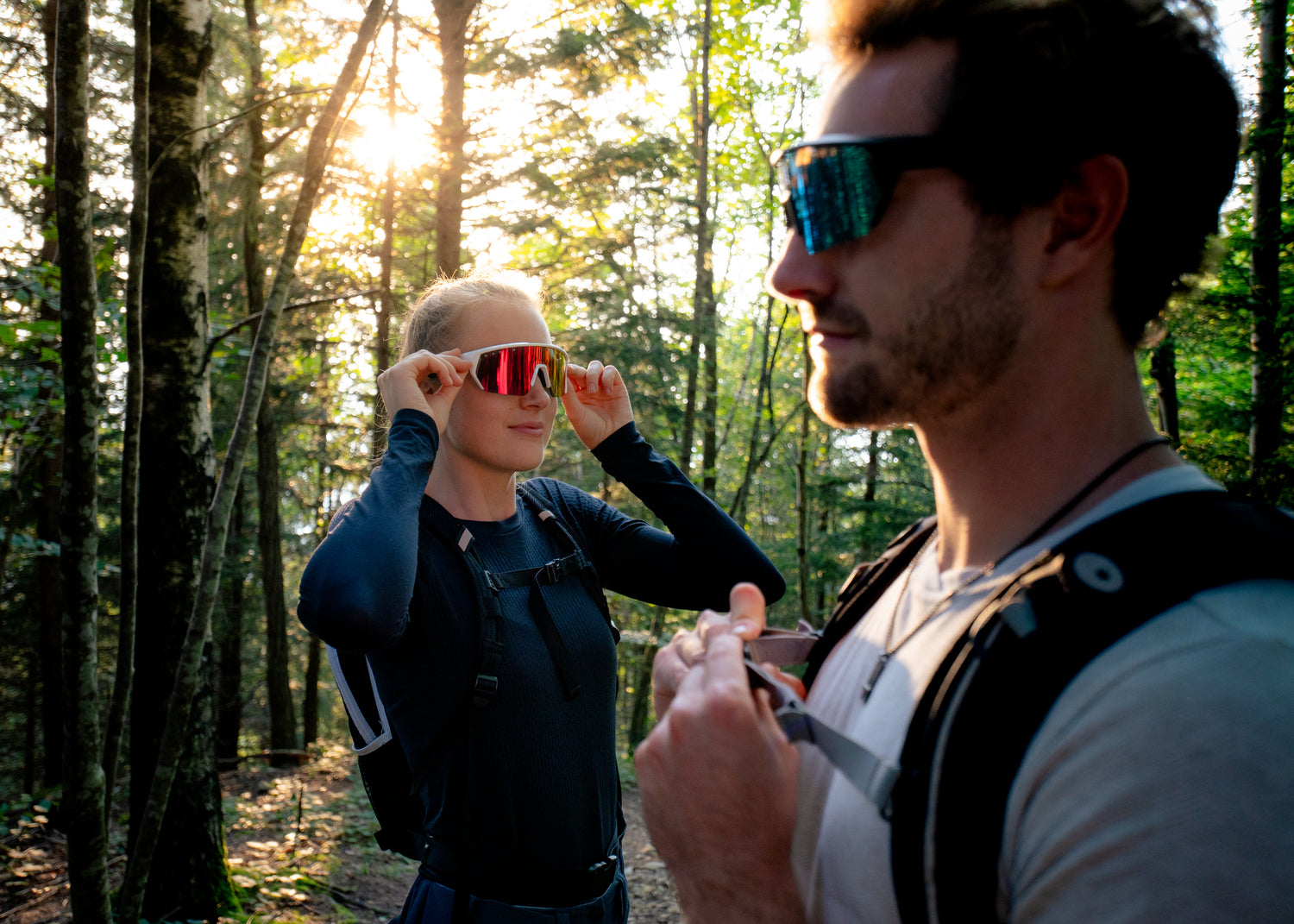 Ein junger Mann und eine junge Frau stehen im Wald, die Frau setzt ihre Brille zum Wandern auf; Credits: Elias Neier