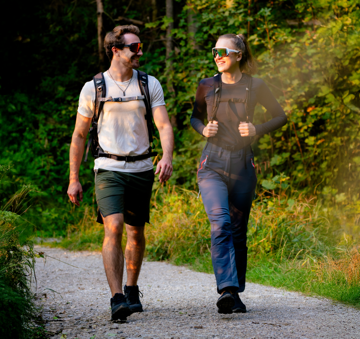 Ein junges Paar wandert auf einer Schottertraße im Wald, sie tragen Wanderrucksäcke und Wanderbrillen; Credits: Elias Neier