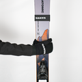 Eine behandschuhte Hand hält einen All-Mountain-Ski mit Bergmotiven fest, auf dem die Markennamen NAKED Optics und Atomic zu sehen sind; der Ski ist mit einem robusten Skigurt (Skifix) gesichert.