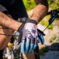 A person wearing The RAID Bike Handschuh by NAKED Optics, featuring a palm tree pattern, rests their hands on mountain bike handlebars outdoors.