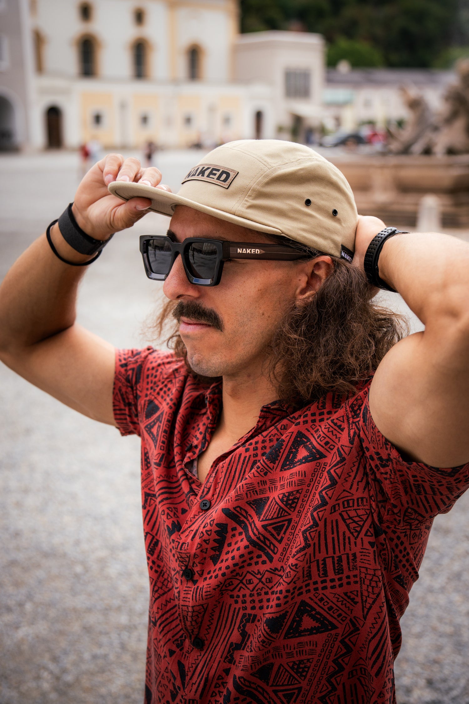 Ein Männ steht in einer Stadt. Er trägt eine Sonnenbrille und setzt sich eine beige Cap auf; Crdits: Elias Neier