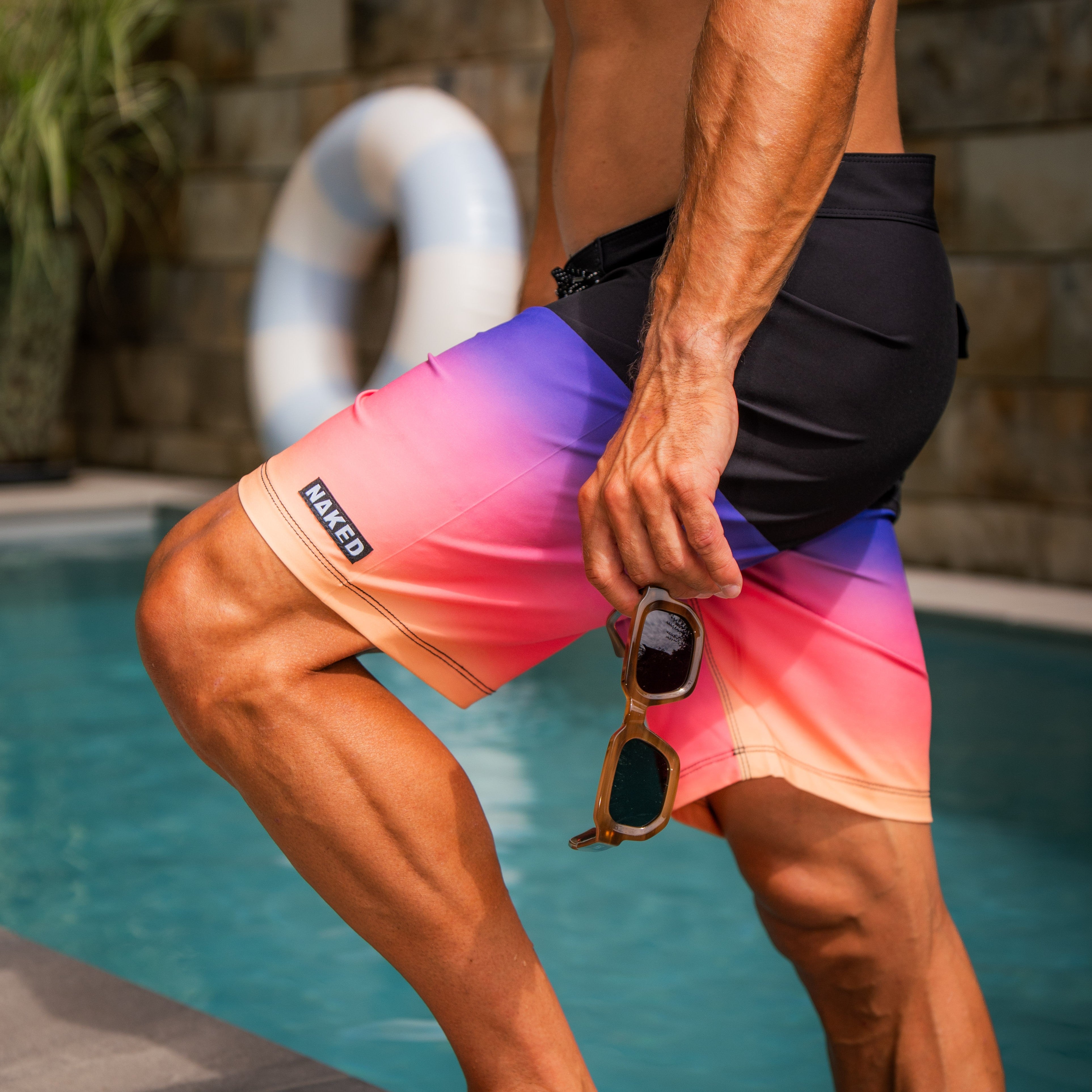 Ein Mann steht am Pool in den „The SHORE“-Boardshorts von NAKED Optics, die aus 4-Wege-Stretchmaterial gefertigt sind – mit einer Sonnenbrille in der Hand, umgeben von üppiger Vegetation und einer Rettungsboje – perfekt vorbereitet zum Surfen oder zum Entspannen in der Sonne.