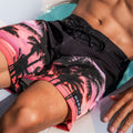 A person relaxing in The SHORE Boardshorts by NAKED Optics, featuring a pink palm tree pattern and crafted from flexible 4-way stretch material, partially submerged in a pool.