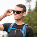 A man wearing NAKED Optics’ The SUMMIT Gletscherbrille with UV400 Schutz and a blue backpack adjusts his glasses outdoors among the trees—prepared for hochalpine Touren.