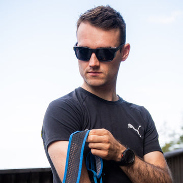 Man in sunglasses by NAKED Optics, wearing The ASCENT Gletscherbrille with UV400 protection and a black Puma shirt, holding a blue strap outdoors on a sunny day—ready for hochalpine Abenteuer or showing off his favorite eyewear.