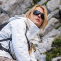 A woman in a white jacket and The SUMMIT Gletscherbrille by NAKED Optics, featuring UV400 Schutz, hiking near rocky terrain on hochalpine Touren and looking up.