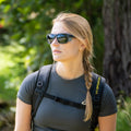 A woman with sunglasses and a braid, wearing a backpack and standing outdoors surrounded by greenery, is ready for alpine Abenteuer with her stylish “The ASCENT” Gletscherbrille from NAKED Optics, offering UV400 Schutz.
