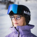 A person in ski gear and glasses stands outdoors in snowy surroundings, wearing a helmet and blue NAKED Optics The TROOP EVO ski goggles with anti-fog coating.