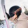 Wearing a tan jacket and helmet with leopard print band, the person sports NAKED Optics’ The STORM skibrille with anti-fog coating as they hold skis on a snowy mountain.