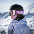 A person in ski gear and a helmet stands in snowy mountains, wearing The TROOP EVO pink ski goggles from NAKED Optics with anti-fog coating and a white jacket.