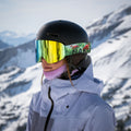 Una donna in tenuta da sci, che indossa gli occhiali da sci “The TROOP EVO” di NAKED Optics con rivestimento antiappannamento e lenti intercambiabili, occhiali riflettenti e un casco a forma di fenicottero, è in piedi su un pendio innevato.