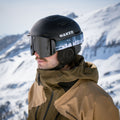 Ein Mann mit einem schwarzen Skihelm und einer NAKED Optics „The TROOP EVO“-Skibrille mit Antibeschlagbeschichtung steht in den verschneiten Bergen und trägt eine braune Jacke.