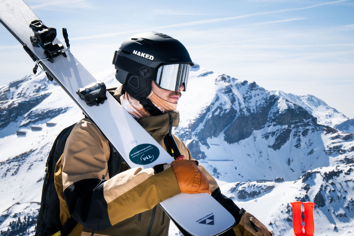 Ein Skifahrer trägt seine Ski auf der Schulter den Berg hinauf. Er trägt einen Skihelm von NAKED Optics, mit der passenden Skibrille; Credits: Elias Neier