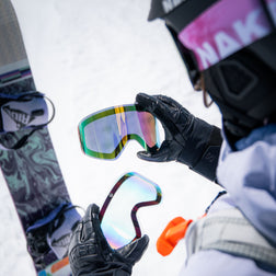 Eine Person in Skikleidung hält mithilfe des innovativen SnapTech-Magnetwechselsystems das Ersatzglas TROOP EVO von NAKED Optics neben einer Skibrille und einem Snowboard auf dem Schnee.