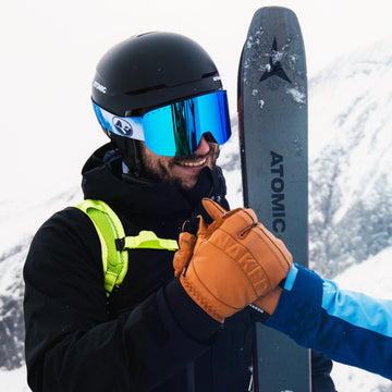 Zwei Skifahrer mit Helmen und Echtleder-Skihandschuhen von NAKED Optics fistbumpen auf einem verschneiten Berg, halten ihre Skier und lächeln - perfekt für Ski- und Snowboard-Fans!.
