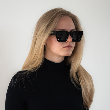 Frau mit langen blonden Haaren, die eine NAKED Optics Sonnenbrille im Retrostil und einen schwarzen Rollkragenpullover trägt und vor einem einfachen Hintergrund posiert.