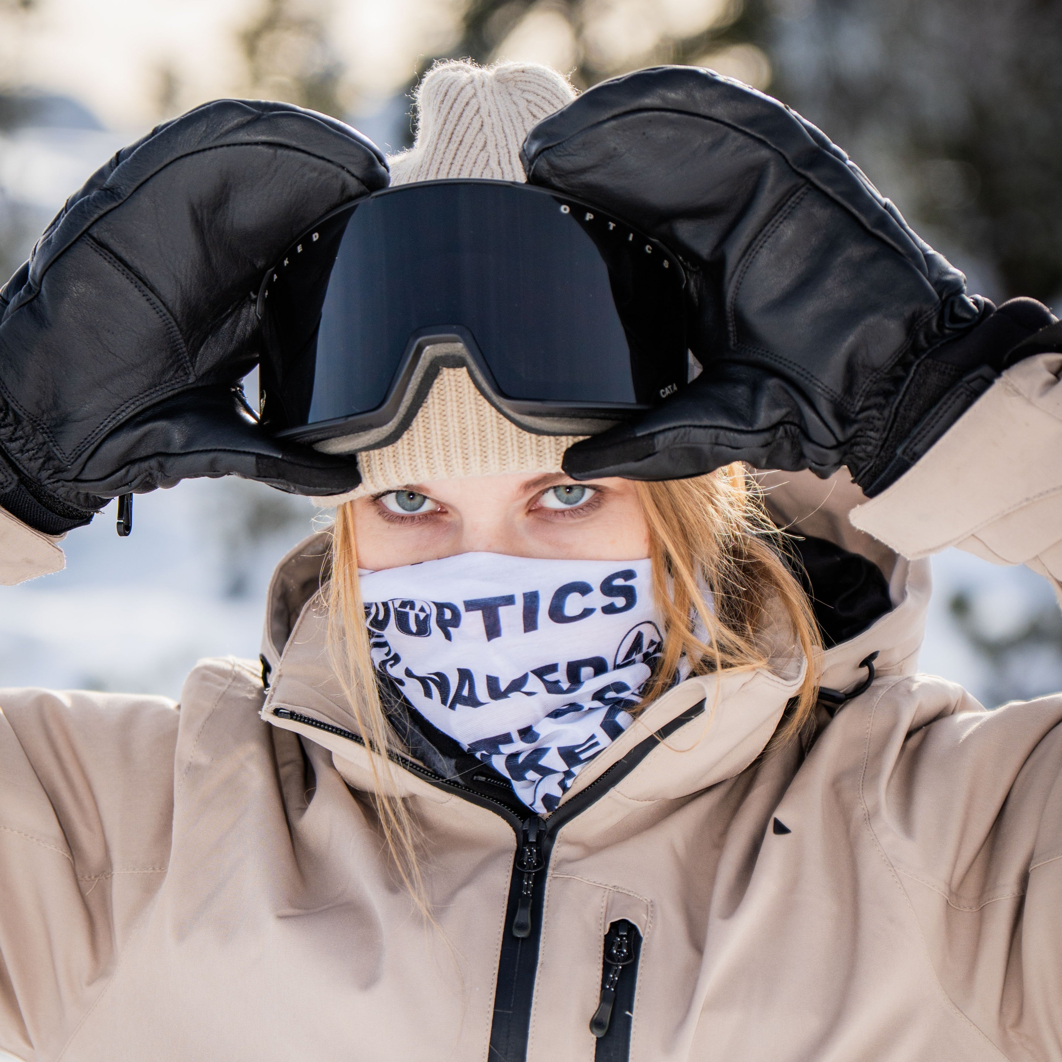 Eine Person in Winterkleidung rückt ihre Skibrille zurecht; sie trägt schwarze Handschuhe, eine hellbraune Jacke und einen gemusterten Schlauchschal (Bandana) von NAKED Optics als Gesichtsbedeckung.