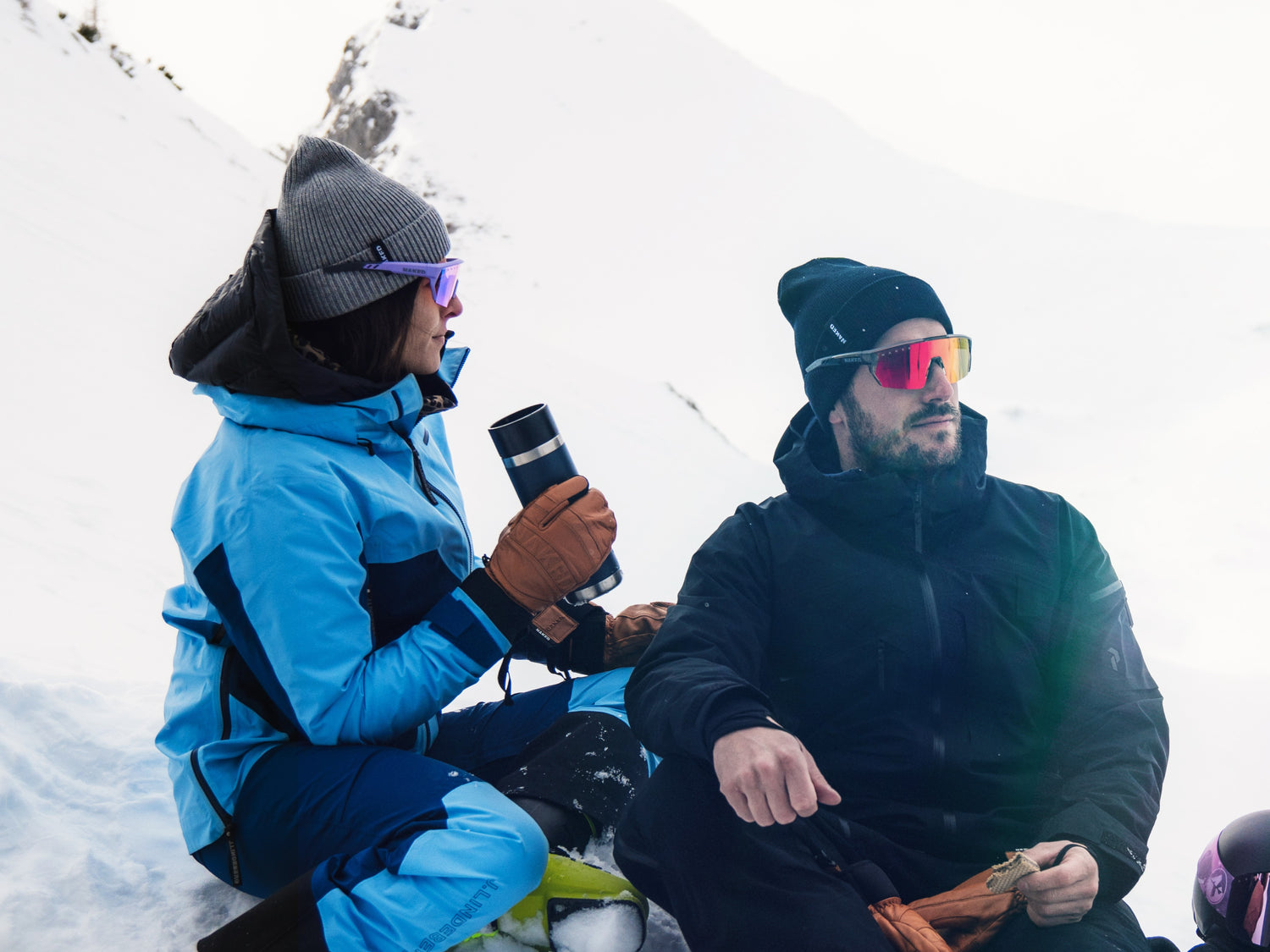 Ein Paar sitz im Schnee und macht eine Pasue, sie tirnken und essen, beide Tragen Beanies und Sportbrillen; Credits: Elias Meier