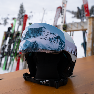 Ein NAKED Optics Skihelm mit Bergmotiv und "NAKED"-Schriftzug, gepaart mit einer NAKED Optics Skibrille, ruht auf einem Holztisch mit Skiern und Schnee im Hintergrund.