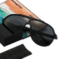 The PILOT sunglasses by NAKED Optics, featuring an oversized design and UV400 dark lenses, rest on their case atop a postcard depicting a stunning beach and ocean scene.