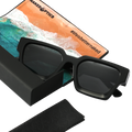 Die schwarze Sonnenbrille The AURA von NAKED Optics mit polarisierten Gläsern liegt neben einem Reinigungstuch mit Strandmotiv und der Aufschrift #lifeisbetternaked – das perfekte Accessoire im angesagten Retro-Design.