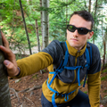Un randonneur portant des lunettes de soleil The RAVE de la marque NAKED Optics et un sac à dos est adossé à un arbre de la forêt.