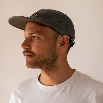 Ein bärtiger Mann im weißen Hemd trägt eine graue 5-Panel NAKED Optics Cap und blickt nach links vor einem schlichten Hintergrund - ideal für Fans von Baumwoll Caps oder stilvollen NAKED Optics Accessoires.