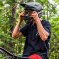 Mountain biker in helmet and goggles adjusts gear while sitting on a bike in a forested area, wearing The RIFT Motocross Brille by NAKED Optics for clear vision on the trails.