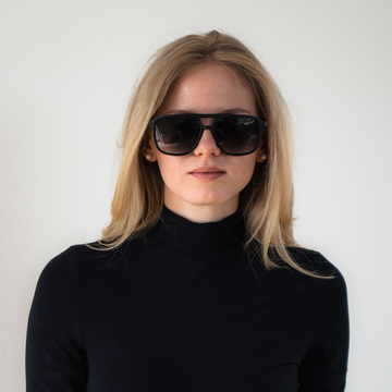Frau mit glattem blondem Haar, die einen schwarzen Rollkragenpullover und eine NAKED Optics-Sonnenbrille im Retrostil mit polarisierten CAT 3-Gläsern trägt, steht vor einem weißen Hintergrund.