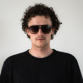A person with curly hair wearing a black shirt and The CARTEL sunglasses by NAKED Optics stands against a plain white background.
